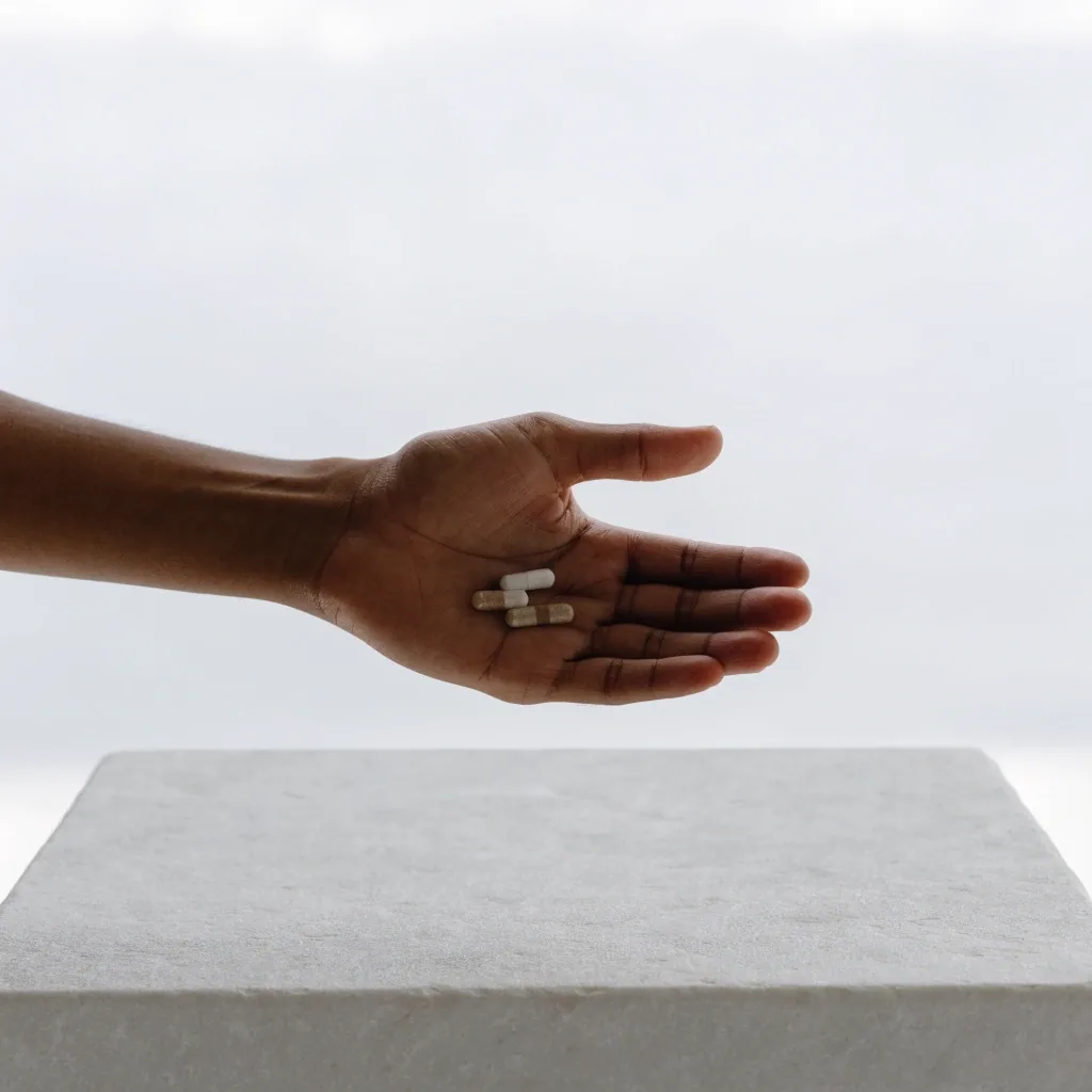 A man's open palm holding three loose supplement capsules over a pale stone bench in flat overcast light