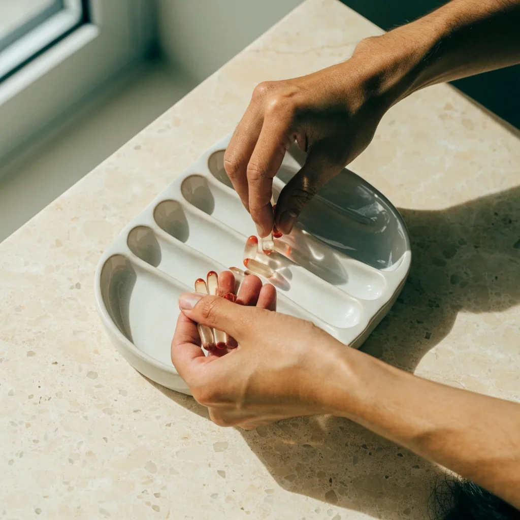A pair of hands sorting matte tablets into the compartments of a shallow pale dish on a granite-look surface