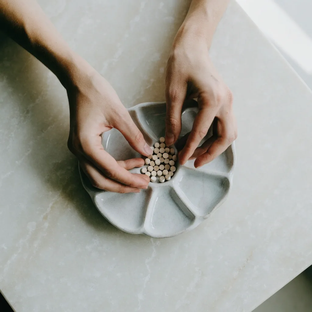 Hands emptying a small quantity of matte tablets into a shallow compartmented dish on a pale bench